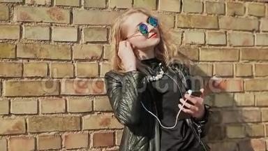 时尚的金发女郎戴着太阳镜和皮夹克，用手机在蓝牙耳机上听音乐
